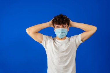 Karantina sırasında Coronavirus virüsü virüsü Cvid-19 'dan korunan 20' li yaşlardaki endişeli genç adam ellerini mavi arka planda izole edilmiş kafaya koydu..