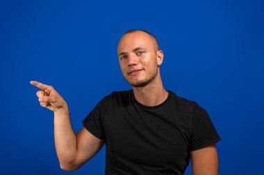 İzole edilmiş mavi arka planda rahat gömlekler giyen genç ve yakışıklı bir adam. Yüzünde mutlu ve doğal bir ifadeyle el ve parmak uçlarını gösteren bir gülümseme var..