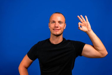 Yüzünde sıradan tişörtler olan neşeli bir adam kameraya gülümsüyor. Tamam işareti mavi arka planda..