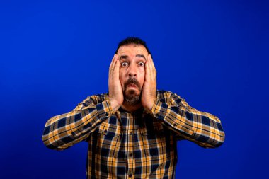 Mavi tek renkli arka planda korkmuş bir duruşu olan genç adam.