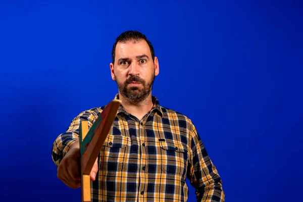 Man in een geel geruit shirt krabt zijn rug met een houten zwaard geïsoleerd op blauwe studio achtergrond. — Stockfoto