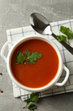 Bir kase domates çorbası, kaşık ve havlu gri arka planda.
