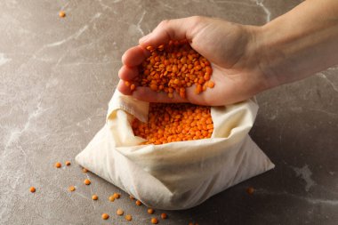 Kadın eli, gri arka plandaki bir çanta dolusu baklagil üzerinde kırmızı baklagilleri tutuyor.