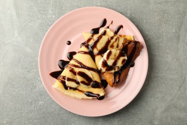 Çikolata soslu krep tabağı ve gri arka planda muz dilimleri.