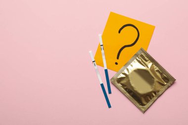 Pembe arka planda prezervatif, soru işareti ve hamilelik testleri