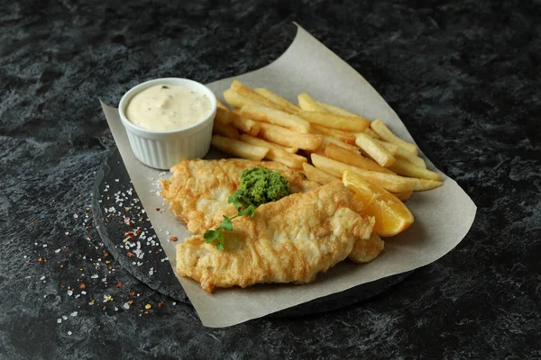 Baking paper with fried fish and chips on black smokey table