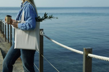 Kağıt bardaklı ve eko çantalı bir kadın denize karşı iskelede duruyor.