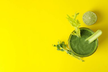 Bir bardak yeşil smoothie ve malzemeler sarı masada.