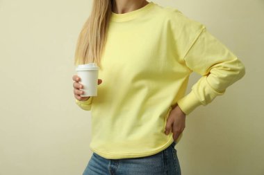 Sarı kazaklı genç kadın bej arka planda kağıt bardak tutuyor.