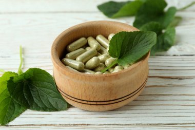 Beyaz ahşap arka planda bitkisel kapsüller ve yapraklarla dolu bir kase.