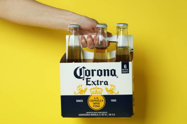 Kadın eli, sarı arka planda Corona Extra ile birlikte karton kutuyu tutuyor.