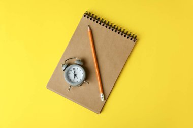 Sarı zemin üzerinde çalar saat, defter ve kalem.