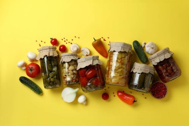Sarı arka planda turşulu sebze kavanozları ve malzemeler.