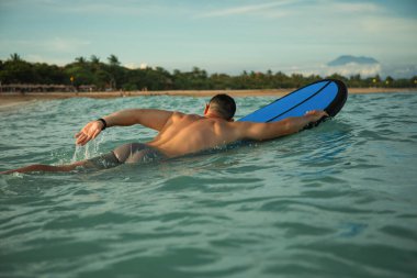 Sörfçü gün batımında sakin sularda uzak dalgalara doğru kürek çeker.
