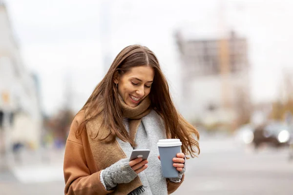 Smiling young woman with smart phone and coffee cup outdoors at urban setting