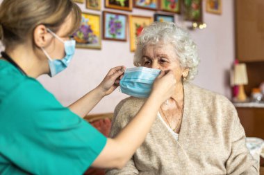 Female nurse helping a senior woman to put on protective face mask during home visit