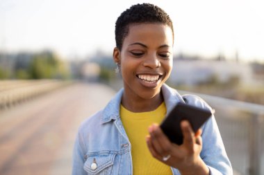 Açık havada akıllı telefon kullanan gülümseyen genç bir kadın.