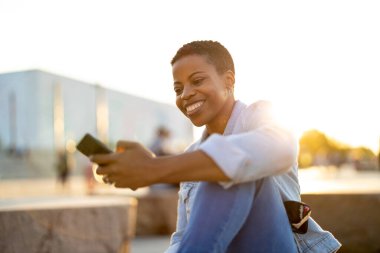 Açık havada akıllı telefon kullanan gülümseyen genç bir kadın.