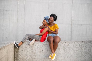 Gri duvarın önünde gülümseyen iki bayan arkadaş.