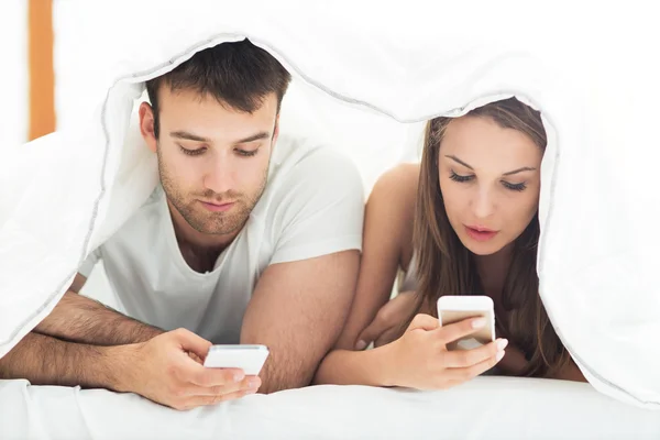 Couple under bed covers with phones — Stock Photo, Image