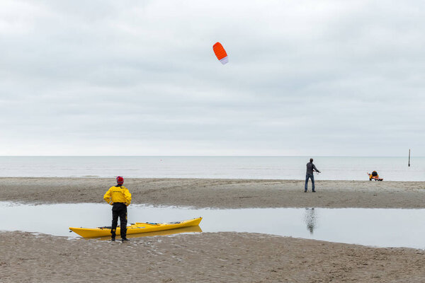 Koksijde, Belgium - November 8, 2020: A man standing next to a yellow kayak, watches another man flying a kite