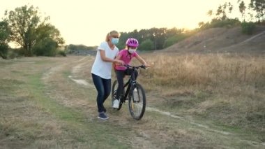Genç bir anne kızına tıbbi maskeli bisiklet sürmeyi ve nehir kenarında bisiklet kaskı takmayı öğretiyor. Covid-19 koronavirüs enfeksiyonundan korunma.