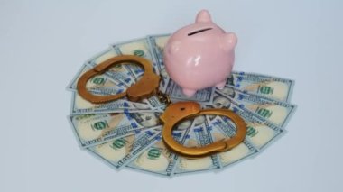 Altın kelepçeler, pembe domuz kumbarası ve beyaz arka planda dönen 100 dolarlık banknotlar. Yolsuzluk ve ekonomik suç kavramı.