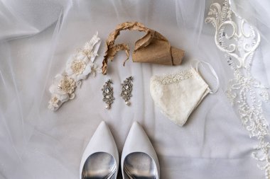Beyaz düğün aksesuarları koleksiyonu içinde duvak küpe, çanta ve beyaz kumaş arka planda düzenlenmiş ayakkabılar da var.