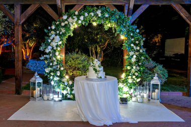 Çiçekli bir kemerin altındaki masada beyaz bir düğün pastası.