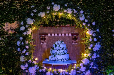 Üzerinde yeşil yapraklar olan beyaz bir pasta, ahşap kemerli bir duvarın önündeki yuvarlak bir masada sergileniyor.