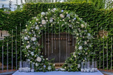 Beyaz çiçekli bir kemer ve zincirlerle süslenmiş bir yeşillik ahşap bir kapının önünde duruyor.