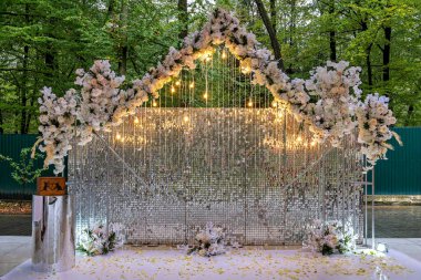 Beyaz çiçek ve ışıklarla süslenmiş beyaz kafes zeminli dekoratif bir yapı parka benzer bir ortamda kurulmuş.