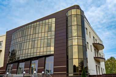 Parçalı bulutlu gökyüzüne karşı yansıtıcı cam ve koyu kahverengi panellerin birleşiminden oluşan kavisli ön cepheli modern bir bina.