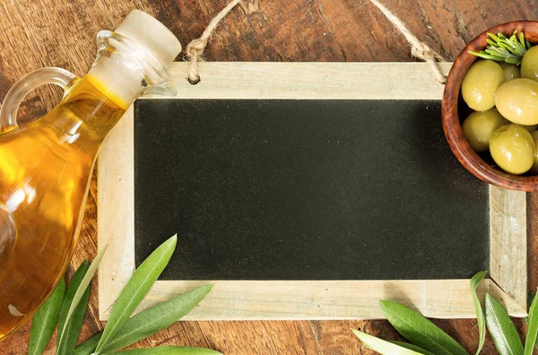 Azeite, azeitonas verdes e folhas sobre quadro-negro sobre mesa de madeira — Fotografia de Stock