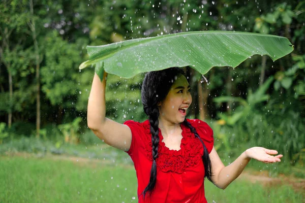 青い庭の背景の雨から避難所を取ってバナナの葉の下に若いアジアの女性の肖像画 — ストック写真