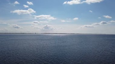 Güneş ışınları suya yansıyor. Gün batımında mavi göl yüzeyinde panoramik manzara dalgalar, orman ve gökyüzü. Gün batımında beyaz bulutlu güzel bir gökyüzü. Doğada vahşi bir yer