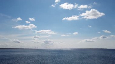 Güneş ışınları suya yansıyor. Gün batımında mavi göl yüzeyinde panoramik manzara dalgalar, orman ve gökyüzü. Gün batımında beyaz bulutlu güzel bir gökyüzü. Doğada vahşi bir yer