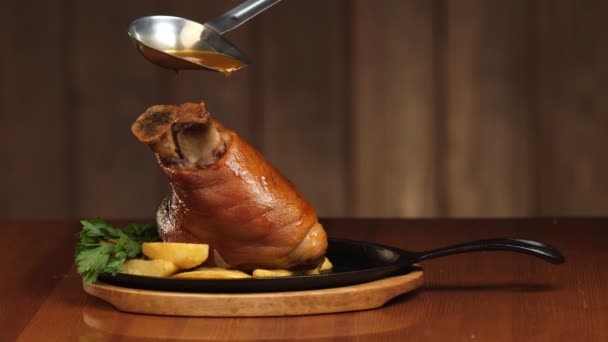 Die Schweinshaxe mit der Sauce aus der Schöpfkelle aufgießen. Fertige Fleischplatte mit Garnitur in einer Pfanne. Knöchel auf einem hölzernen Hintergrund. Zeitlupe — Stockvideo