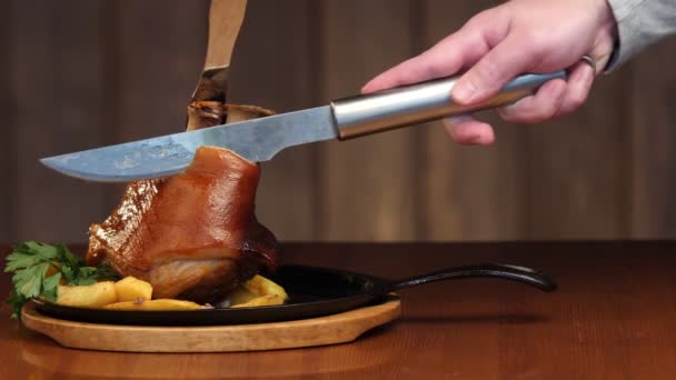 Een mensenhand snijdt een groot stuk vlees van een varkensknokkel met een mes. Kant en klare vleesschotel met garnering in een koekenpan. Knuffel op een houten ondergrond. Langzame beweging — Stockvideo