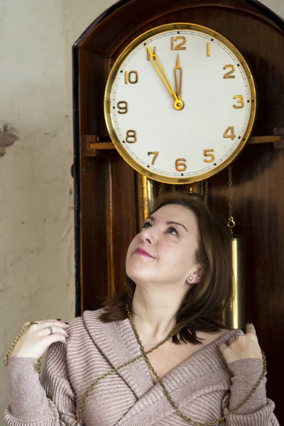 A brunette next to a large grandfather clock. Holding time. Women\'s time.