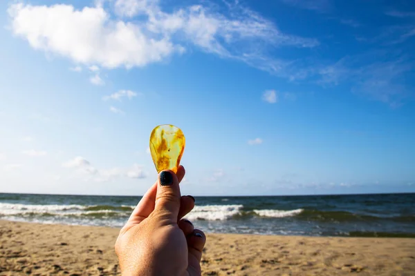 Ambra. Molto bella ambra trasparente nel palmo della mano. Sulla spiaggia sabbiosa si trova una mano con una pietra ambrata. Luminosa e trasparente ambra contro un cielo azzurro brillante. Resina di legno congelata. Baltico — Foto Stock