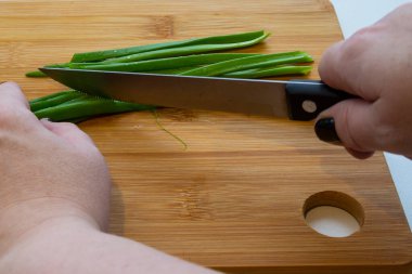 Bıçaklı bir kadın eli, beyaz bir arka planda tahta bir tahtada sebzeleri dilimliyor. Kadın yeşil soğan kesiyor.