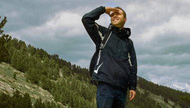 Yürüyüş ve ileriye dönük arka planda yeşil orman ile tepenin tepesinde duran sırt çantası ile sağlıklı genç gülümseyen adam Portresi - Sucess kavramı