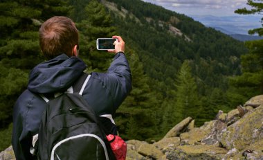 Genç bir gezgin adam Prespa Milli Parkı'nda dağın tepesinde oturup akıllı telefon ile manzara resimleri çekmek