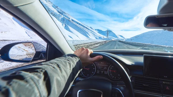 Snowy mountains road view from the modern luxury car interior with drivers hand on steering wheel — Stock Fotó