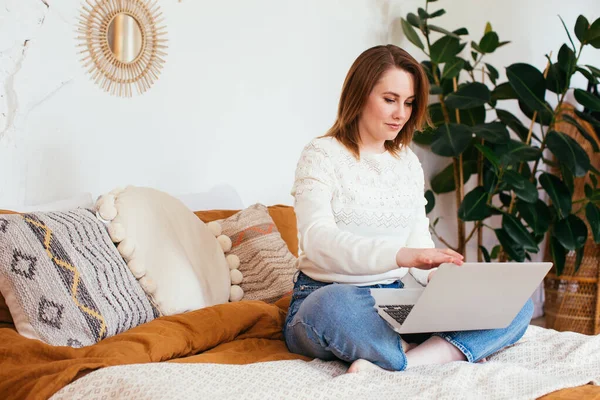 Pretty smiling woman has pleasant smile, haircut, dressed in casual domestic clothes, laugh during watching comedy, leaves comments chats online with friends on laptop computer sits in bed. — Stock Photo, Image