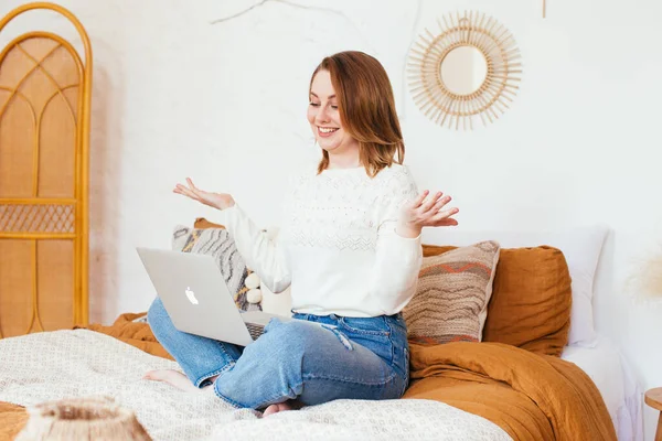 Pretty smiling woman has pleasant smile, haircut, dressed in casual domestic clothes, laugh during watching comedy, leaves comments chats online with friends on laptop computer sits in bed. — Stock Photo, Image