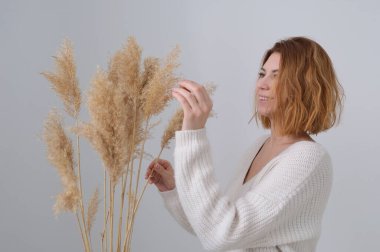 Kızıl saçlı kadın otları elinde tutuyordu. Reed sapı, kurutulmuş otlar, ev için dekoratif tüy kompozisyonu, plaj teması, yeni moda ev dekorasyonu.