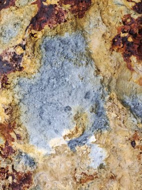 Yıpranmış bir kaya yüzeyinin canlı bir renk kontrastını gösteren makro yakın çekim görüntüsü. Kompozisyonda soluk mavi-gri mineralden oluşan merkezi bir yama yer alıyor.