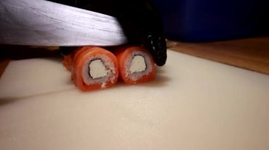 Siyah lastik eldivenli aşçı. Suşi rulosunu taze somon balığı ve krem peynirle kes. Keskin bir bıçakla beyaz bir tahta üzerinde. Philadelphia suşisi hazırlanıyor. Japon mutfağı..